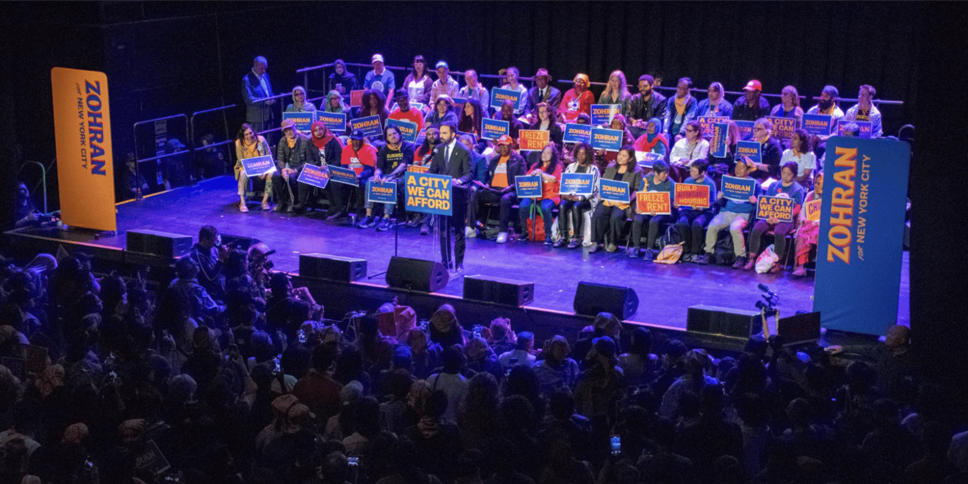 Zohran Mamdani stands at a podium in front of a crowd of supporters who hold signs. Zohran Mamdani stands at a podium in front of a crowd of supporters who hold signs.